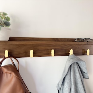 May include: A wooden wall shelf with a dark brown finish, featuring four gold-colored hooks. A small potted plant and eyeglasses rest on the shelf. A brown backpack and a gray jacket hang from the hooks. The shelf is mounted on a white wall.