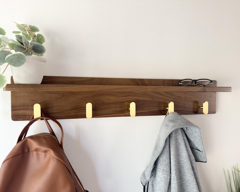 May include: A wooden wall shelf with a dark brown finish, featuring four gold-colored hooks. A small potted plant and eyeglasses rest on the shelf. A brown backpack and a gray jacket hang from the hooks. The shelf is mounted on a white wall.