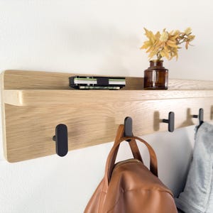 May include: A wooden wall shelf with a light brown finish, featuring black hooks and a top shelf. A brown leather bag and a gray jacket hang from the hooks. A small brown vase with dried flowers and a wallet rest on the shelf.