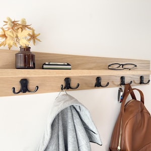 May include: A wooden wall-mounted shelf with a built-in shelf and five black metal hooks. A brown leather backpack, a gray jacket, and keys hang from the hooks. The shelf holds a small vase with dried flowers, glasses, and a small tray.