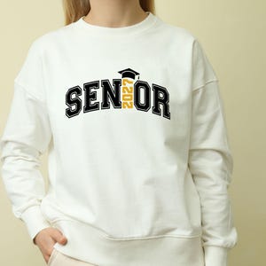 Puede incluir: Una sudadera blanca de cuello redondo con el texto negro SENIOR, donde el n&uacute;mero 2027 en amarillo reemplaza la letra I. Un icono de birrete de graduaci&oacute;n negro se sit&uacute;a sobre el n&uacute;mero 2027. El dise&ntilde;o est&aacute; centrado en el pecho de la prenda.