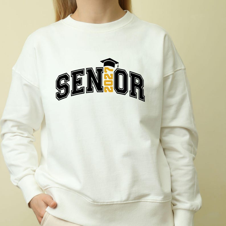 Puede incluir: Una sudadera blanca de cuello redondo con el texto negro SENIOR, donde el n&uacute;mero 2027 en amarillo reemplaza la letra I. Un icono de birrete de graduaci&oacute;n negro se sit&uacute;a sobre el n&uacute;mero 2027. El dise&ntilde;o est&aacute; centrado en el pecho de la prenda.