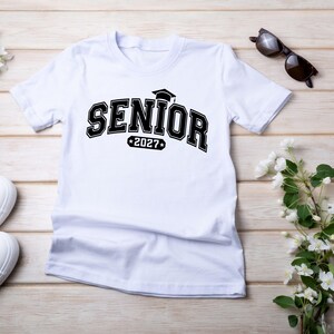 Puede incluir: Una camiseta blanca de manga corta con el texto negro SENIOR en una fuente universitaria en negrita, con un icono de birrete de graduaci&oacute;n encima y el a&ntilde;o 2027 dentro de un borde rectangular con estrellas a los lados.