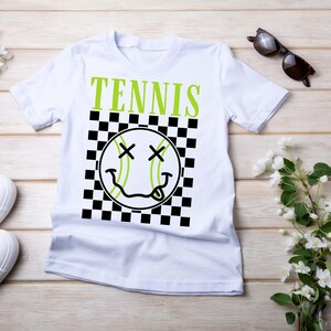 May include: White t-shirt with a black and white checkered pattern and a green tennis ball with a smiley face. The text "TENNIS" is printed in green above the ball.