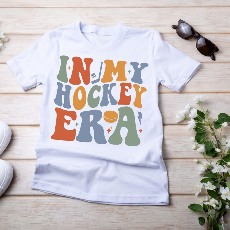 K&ouml;nnte beinhalten: Wei&szlig;es T-Shirt mit dem Text "IN MY HOCKEY ERA" in Retro-Schrift. Das Design beinhaltet einen Hockeyschl&auml;ger, einen Puck und Sterne in verschiedenen Farben. Sonnenbrille und Blumen liegen auf einem Holzgrund.