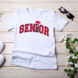 Puede incluir: Una camiseta blanca de manga corta con el texto SENIOR en rojo y negro, con el a&ntilde;o 2027 integrado verticalmente en el centro, rematado con un icono de birrete de graduaci&oacute;n rojo. El dise&ntilde;o est&aacute; impreso en una fuente de bloque universitaria.