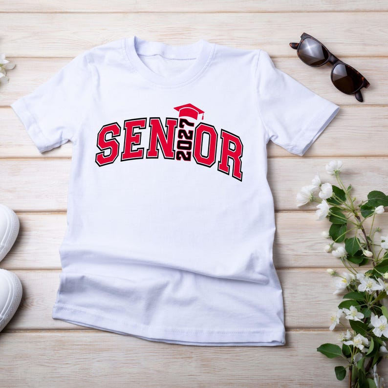 Puede incluir: Una camiseta blanca de manga corta con el texto SENIOR en rojo y negro, con el a&ntilde;o 2027 integrado verticalmente en el centro, rematado con un icono de birrete de graduaci&oacute;n rojo. El dise&ntilde;o est&aacute; impreso en una fuente de bloque universitaria.