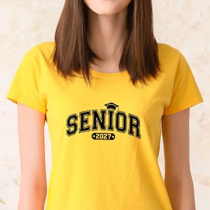Puede incluir: Una camiseta de manga corta de color amarillo brillante con la palabra SENIOR en letras negras gruesas de estilo universitario. Un peque&ntilde;o icono de birrete negro se sit&uacute;a sobre el texto y el n&uacute;mero 2027 aparece impreso en un borde rectangular.