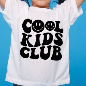 May include: White t-shirt with black text that reads "Cool Kids Club" in a retro font. Two smiley faces are above the text.