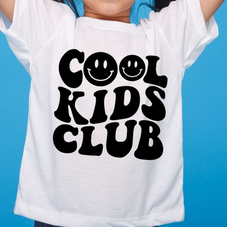 May include: White t-shirt with black text that reads "Cool Kids Club" in a retro font. Two smiley faces are above the text.