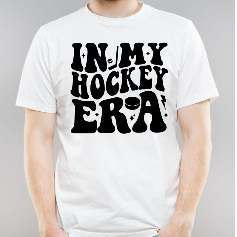 K&ouml;nnte beinhalten: Wei&szlig;es T-Shirt mit dem Text "IN MY HOCKEY ERA" in fetter, schwarzer Retro-Schrift. Das Design beinhaltet Hockey-Grafiken wie Schl&auml;ger und Puck. Das T-Shirt ist f&uuml;r den l&auml;ssigen Gebrauch bestimmt.