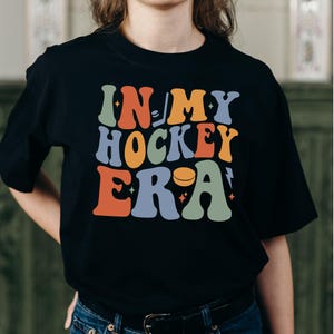 K&ouml;nnte beinhalten: Schwarzes T-Shirt mit dem Text "IN MY HOCKEY ERA" in Retro-Schrift. Der Text ist in verschiedenen Farben gehalten, mit Hockey-Grafiken. Ein l&auml;ssiges Kleidungsst&uuml;ck.