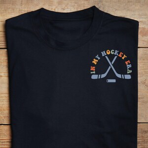 K&ouml;nnte beinhalten: Marineblaues T-Shirt mit dem Text "IN MY HOCKEY ERA" in einem Halbkreis &uuml;ber gekreuzten Hockeyschl&auml;gern und einem Puck. Der Text und die Grafik sind in Orange-, Blau- und Gelbt&ouml;nen gehalten.