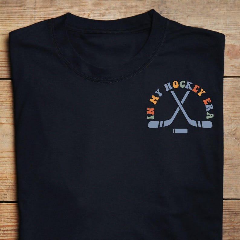 K&ouml;nnte beinhalten: Marineblaues T-Shirt mit dem Text "IN MY HOCKEY ERA" in einem Halbkreis &uuml;ber gekreuzten Hockeyschl&auml;gern und einem Puck. Der Text und die Grafik sind in Orange-, Blau- und Gelbt&ouml;nen gehalten.