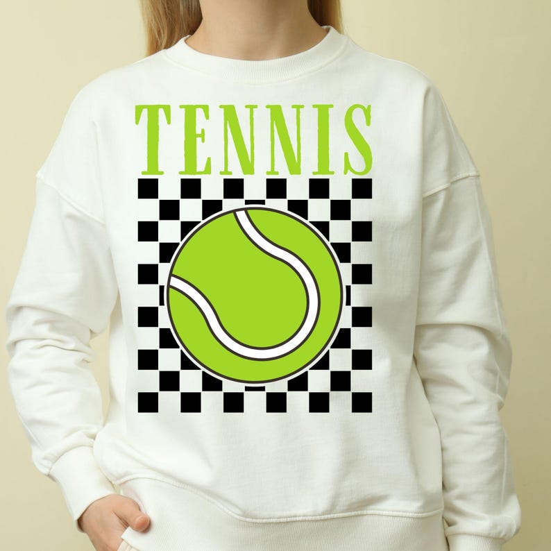 May include: White sweatshirt with the word "TENNIS" printed in green letters. A green tennis ball with a white seam is centered on a black and white checkered background.
