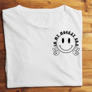 K&ouml;nnte beinhalten: Wei&szlig;es T-Shirt mit einem schwarzen Grafik-Smiley, der mit beiden H&auml;nden ein Friedenszeichen macht. Der Text "IN MY HOCKEY ERA" ist um das Smiley herum gedruckt. Das T-Shirt liegt flach auf einer Holzoberfl&auml;che.