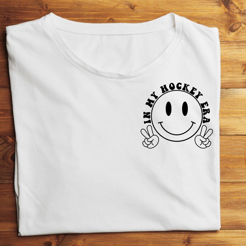 K&ouml;nnte beinhalten: Wei&szlig;es T-Shirt mit einem schwarzen Grafik-Smiley, der mit beiden H&auml;nden ein Friedenszeichen macht. Der Text "IN MY HOCKEY ERA" ist um das Smiley herum gedruckt. Das T-Shirt liegt flach auf einer Holzoberfl&auml;che.