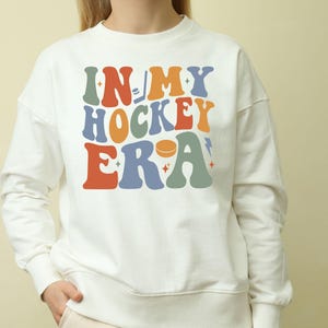 K&ouml;nnte beinhalten: Ein wei&szlig;es Sweatshirt mit dem Text "IN MY HOCKEY ERA" in bunten Buchstaben im Retro-Stil. Das Design beinhaltet einen Hockeypuck und eine Musiknote. Ein l&auml;ssiges Kleidungsst&uuml;ck.
