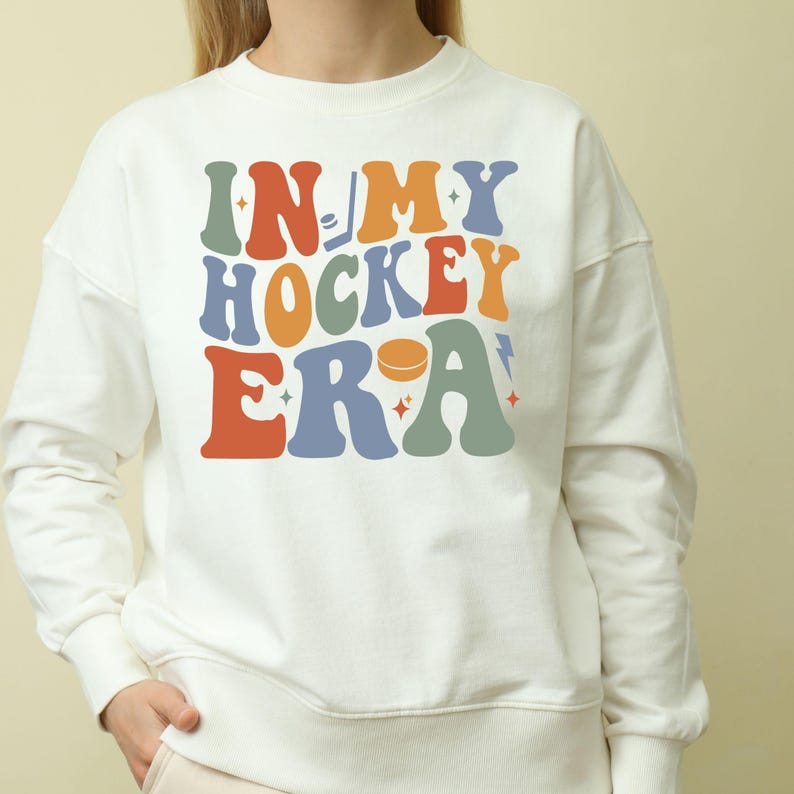 K&ouml;nnte beinhalten: Ein wei&szlig;es Sweatshirt mit dem Text "IN MY HOCKEY ERA" in bunten Buchstaben im Retro-Stil. Das Design beinhaltet einen Hockeypuck und eine Musiknote. Ein l&auml;ssiges Kleidungsst&uuml;ck.