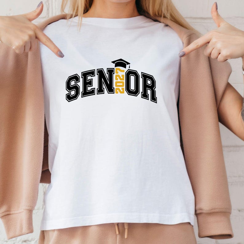 Puede incluir: Una camiseta blanca con el texto negro SENIOR en una fuente universitaria en negrita. La letra I est&aacute; reemplazada por el n&uacute;mero amarillo 2027 y un icono de birrete de graduaci&oacute;n negro se sit&uacute;a justo encima del n&uacute;mero, centrado en el pecho.