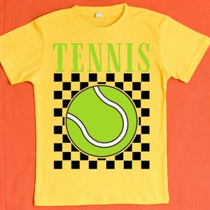 May include: Yellow t-shirt with a black and white checkered pattern and a green tennis ball graphic. The text "TENNIS" is printed in black letters above the graphic.