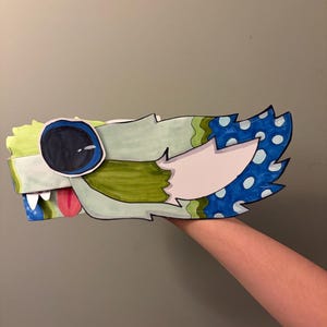 Puede incluir: Una cabeza de dragón de papel hecha a mano, con un ala de lunares azules y blancos. La cabeza del dragón presenta un gran ojo negro, una lengua rosa y detalles en verde, azul y blanco. La obra de arte está sostenida por una mano.