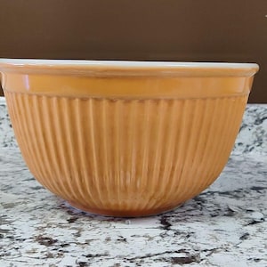 May include: A round mixing bowl with a glossy orange exterior and a white interior. The bowl features a distinct vertical ribbed texture around the entire outer surface, creating a series of parallel lines that extend from the rim to the base.