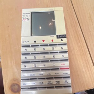 以下が含まれることがあります： ベージュ色のSaitek Pro Bridge 200電子携帯ゲーム機。長方形の画面、黒いロゴ、トランプのマーク、数字、Deal、Score、View Handsなどの操作ボタンを備えたキーボードがあり、白いプラスチック製の筐体で構成されています。