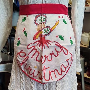 May include: A white apron with a red waistband featuring a hand-painted illustration of a figure in a hat and muff, surrounded by green holly leaves and red berries. The text Merry Christmas is written in red cursive script across the bottom.