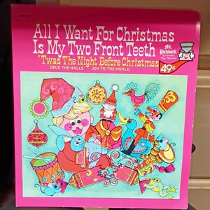 May include: A vintage Mr. Pickwick vinyl record sleeve with a bright pink border. The cover features a colorful illustration of a child with two front teeth missing, surrounded by Christmas toys, ornaments, and animals on a light blue background.