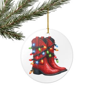 May include: A round Christmas ornament featuring a pair of red cowboy boots wrapped in colorful Christmas lights and blue star accents. The boots have decorative stitching and are hanging from a gold string.