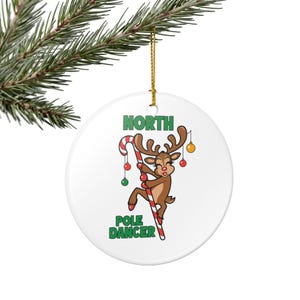 Könnte beinhalten: Weißer Keramik-Weihnachtsschmuck mit einem Cartoon-Rentier, das eine Zuckerstange hält. Das braune Rentier hat ein Geweih und ist mit kleinen Kugeln verziert. Der Text "NORTH POLE DANCER" ist auf dem Ornament aufgedruckt.