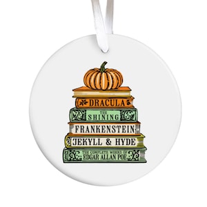 Könnte beinhalten: Ein weißer Keramik-Anhänger mit einem Stapel Bücher mit klassischen Horror-Titeln, darunter "Dracula", "Shining", "Frankenstein", "Jekyll & Hyde" und "The Complete Works of Edgar Allan Poe". Auf den Büchern liegt ein orangefarbener Kürbis.