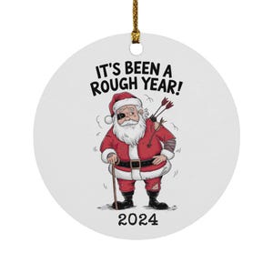 May include: A white round Christmas ornament with a cartoon illustration of Santa Claus with a bandaged arm and an eyepatch. He is holding a cane and has arrows stuck in his back. The text "It's been a rough year!" is above him and the year "2024" is below him.