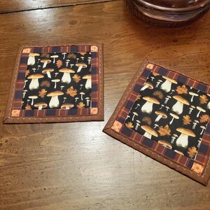 May include: Two square brown and black fabric coasters with a mushroom and leaf pattern. The coasters have a brown border with a scalloped edge.