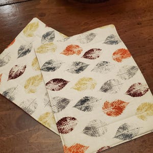 May include: Two off-white placemats with a leaf pattern in shades of orange, yellow, gray, and burgundy. The placemats are rectangular and appear to be made of fabric. The background is a dark wooden surface.