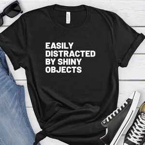 Puede incluir: Camiseta negra con texto blanco que dice "Easily Distracted By Shiny Objects". La camiseta está colocada sobre una superficie de madera con un par de vaqueros y zapatillas negras.