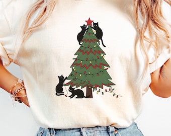 May include: Ivory-colored t-shirt featuring a Christmas tree decorated with lights and a red star. Black cats are climbing and sitting around the tree. The word "IVORY" is printed at the bottom of the image.