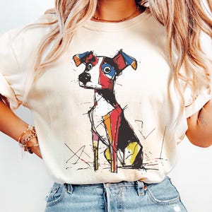 Puede incluir: Camiseta de color marfil con una ilustración geométrica de un perro colorido. El diseño del perro incorpora formas rojas, azules, amarillas y negras. La camiseta tiene mangas cortas y un corte holgado. La palabra "IVORY" está impresa en la parte inferior.