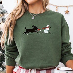 Puede incluir: Sudadera verde militar con un gr&aacute;fico de un perro negro persiguiendo a un mu&ntilde;eco de nieve. El perro lleva un juguete rojo en la boca. El mu&ntilde;eco de nieve lleva una bufanda roja. El texto "MILITARY GREEN" est&aacute; en la parte inferior. Tambi&eacute;n se ve una taza blanca con un dise&ntilde;o de Pap&aacute; Noel.