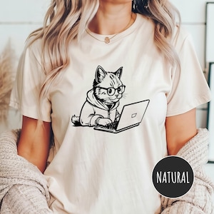 May include: A white t-shirt with a black and white illustration of a cat wearing glasses and using a laptop. The text "NATURAL" is printed on a black circle in the bottom right corner of the image.