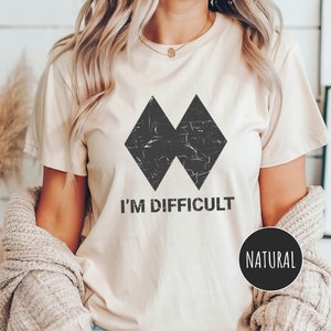 May include: A white t-shirt with a black diamond graphic and the text "I'm Difficult". The shirt is a natural colour.