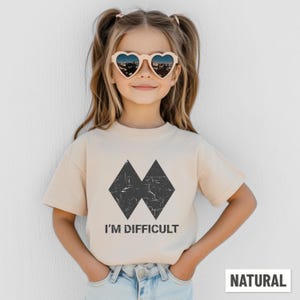 May include: A beige t-shirt featuring a black graphic of two overlapping diamond shapes and the text "I'M DIFFICULT". The shirt is labelled "NATURAL" and is worn by a person wearing heart-shaped sunglasses.