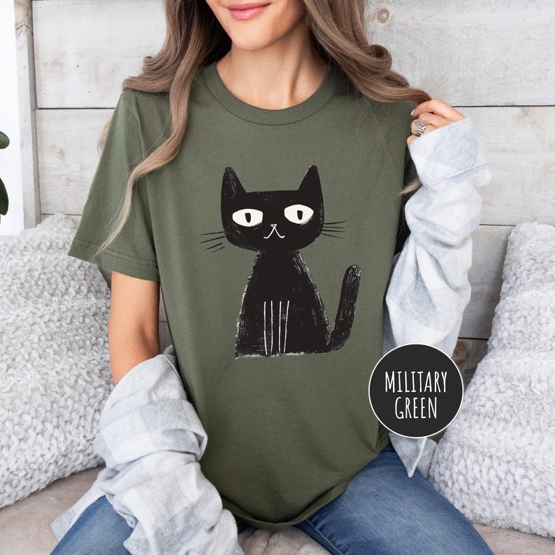 Pu&ograve; includere: Una donna indossa una maglietta verde militare con un'immagine di un gatto nero. Il gatto ha gli occhi bianchi e la bocca bianca. Il testo "Verde Militare" &egrave; su un cerchio nero nell'angolo in basso a destra dell'immagine.