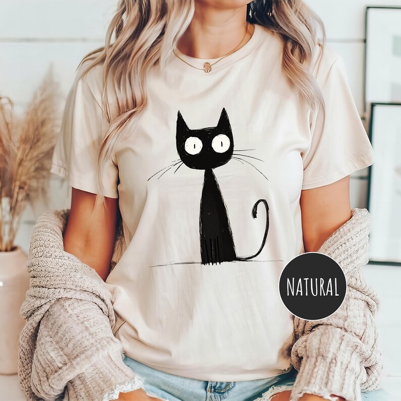 May include: A white t-shirt with a black cat illustration. The cat has large, round eyes and a long tail. The word "NATURAL" is printed on the bottom right corner of the shirt.