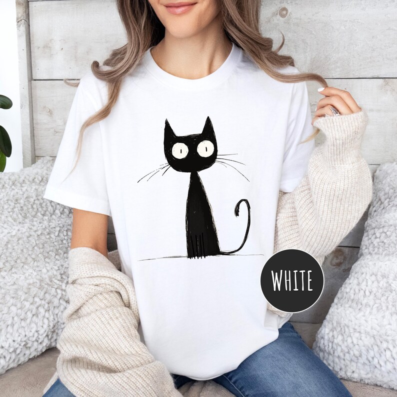 May include: A white t-shirt with a black cat illustration. The cat has large eyes and a long tail. The text "WHITE" is visible on the bottom right corner of the image.