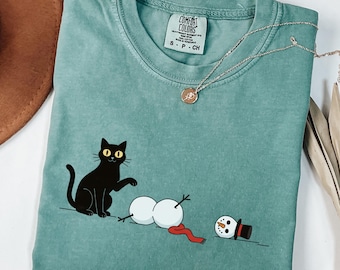Puede incluir: Camiseta verde claro Comfort Colors con un diseño de gato negro y muñeco de nieve. El muñeco de nieve está en pedazos con una bufanda roja y un sombrero de copa negro. El texto "LIGHT GREEN" está en la parte inferior.