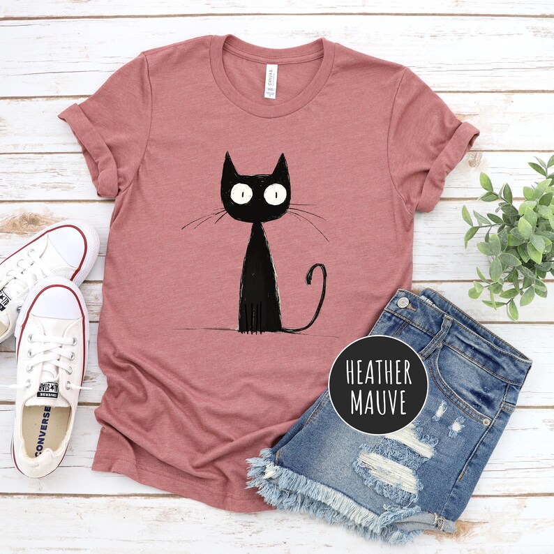 May include: A heather mauve t-shirt with a black cartoon cat graphic. The cat has large eyes and a long tail.