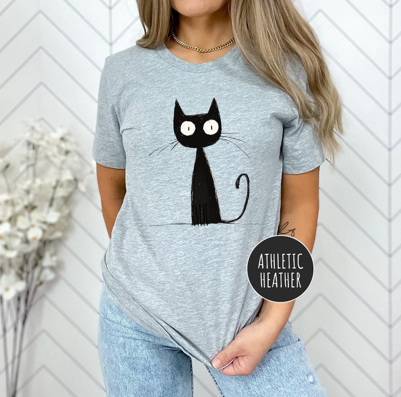 May include: A gray t-shirt with a black cat illustration. The cat has large eyes and a long tail. The text "Athletic Heather" is printed on the bottom right corner of the shirt.