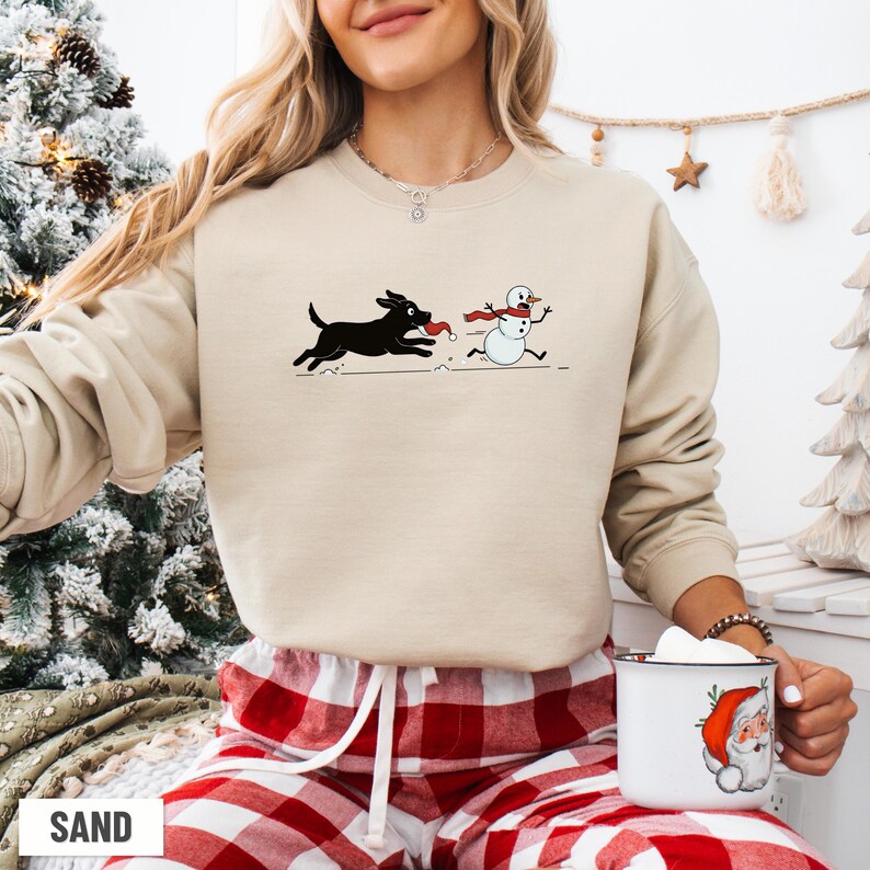 Puede incluir: Sudadera de color arena con un perro negro persiguiendo a un mu&ntilde;eco de nieve con una bufanda roja. El dise&ntilde;o gr&aacute;fico es una escena invernal l&uacute;dica. La sudadera est&aacute; hecha de un material suave.